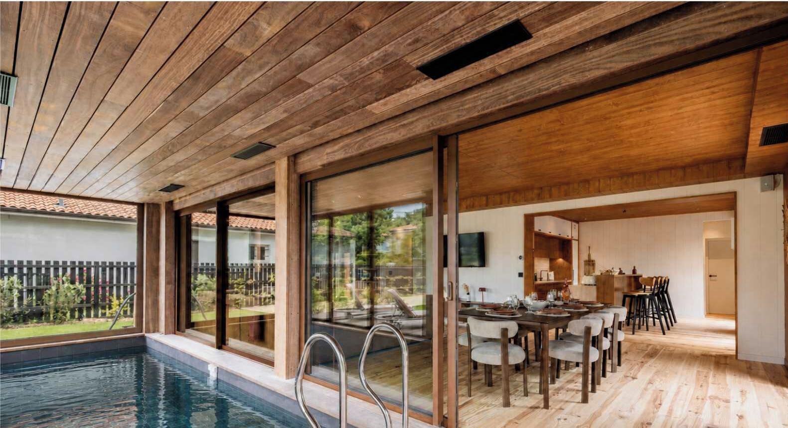 A modern indoor pool area with a wooden ceiling, a dining area with a table and chairs, and large windows offering a view of the outside.