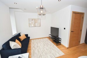 A living room with a dark blue sofa, a flat-screen TV on a stand, a patterned rug, and a wooden door. - Contemporary 1 Bed Apartment in Central Leeds (Leeds)