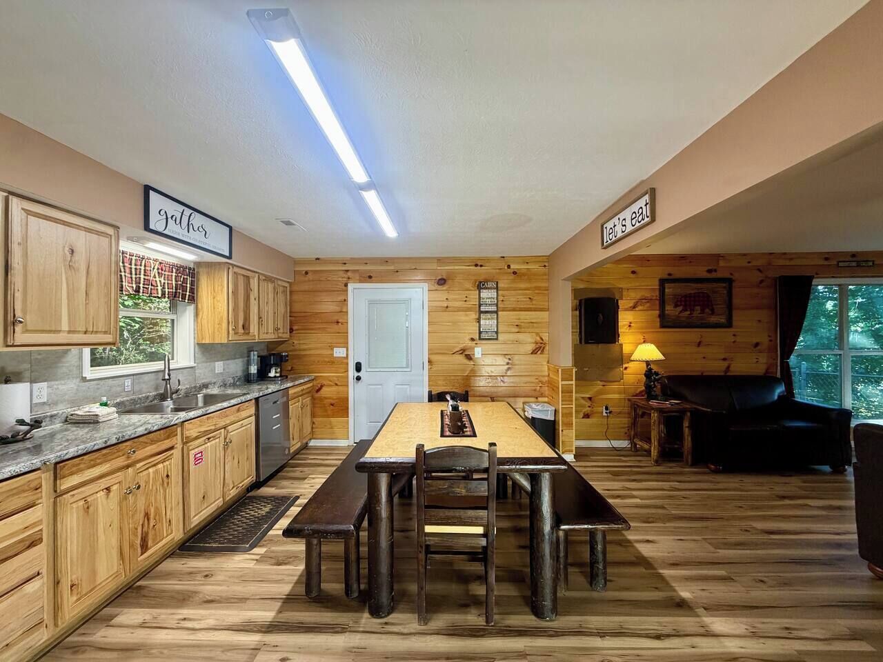 Un intérieur de style cabane avec une table à manger en bois, des bancs, un coin cuisine doté de meubles en bois et un espace salon confortable avec une lampe.