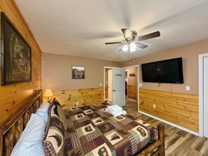 Een slaapkamer met een houten hoofdbord, een gewatteerde spreideken, een plafondventilator, een aan de muur bevestigde televisie en een deur naar een andere kamer.