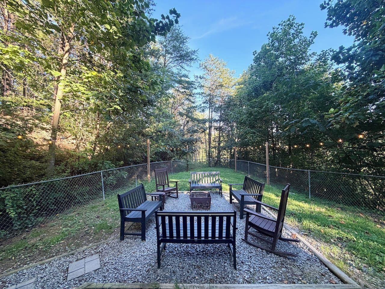 An outdoor seating area with benches and a fire pit, surrounded by a fence and trees.