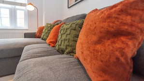Un sofá gris con varios cojines de diferentes colores, una lámpara de pie y una ventana con persianas.