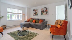 Una sala de estar con un sofá seccional gris, una mesa de centro redonda y un sillón naranja. Hay dos cuadros abstractos en la pared y una ventana con persianas.