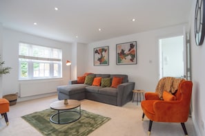 Ein Wohnzimmer mit einem grauen Ecksofa, einem runden Couchtisch und einem orangefarbenen Sessel. An der Wand hängen zwei abstrakte Gemälde und ein Fenster mit Jalousien.