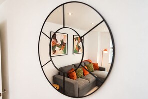 Un miroir rond reflète un salon avec un canapé gris, des coussins colorés et une œuvre d’art abstraite accrochée au mur.