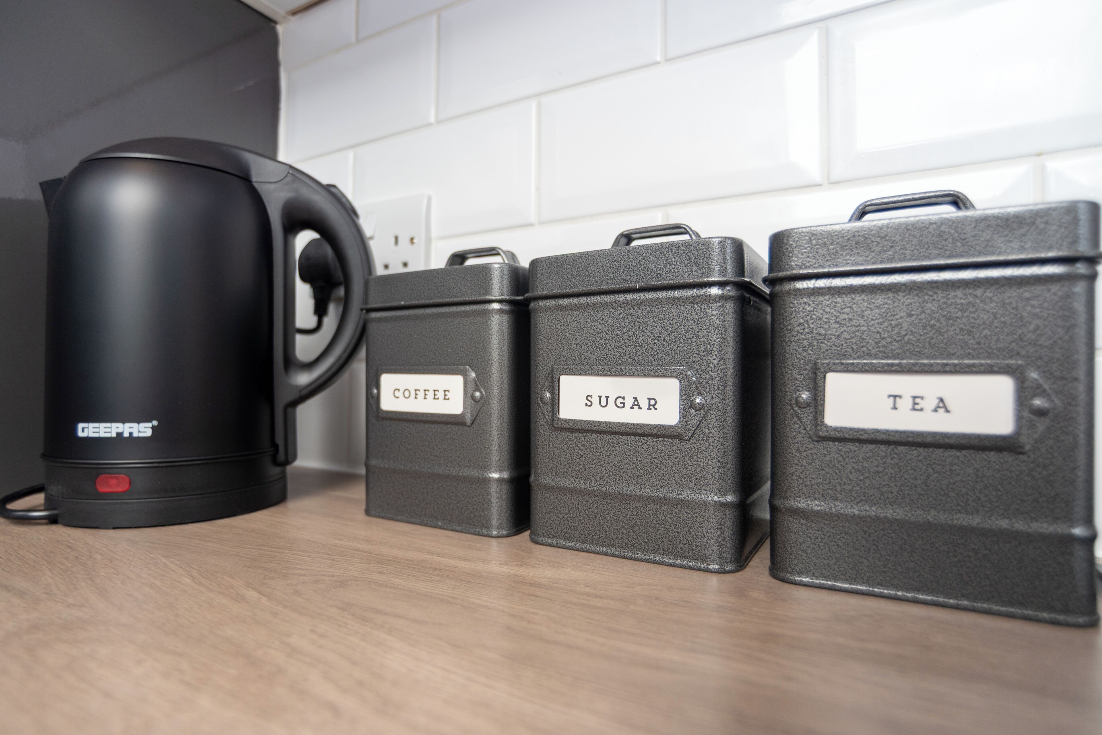 Three dark-colored storage containers with labels on a kitchen counter, one of which is labeled 'COFFEE'.