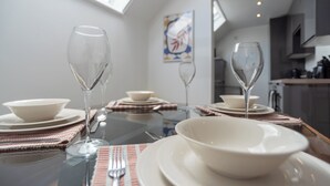 Una mesa de comedor dispuesta con platos blancos, tazones y copas de cristal, con una cocina al fondo.