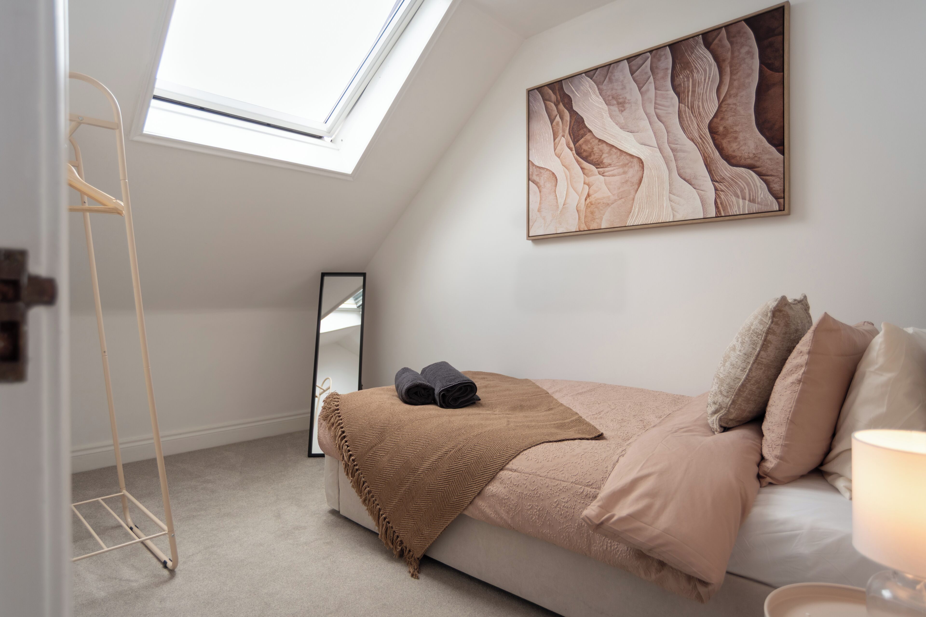 A bedroom with a sloped ceiling, a bed with a brown blanket, a large mirror, and a painting on the wall.