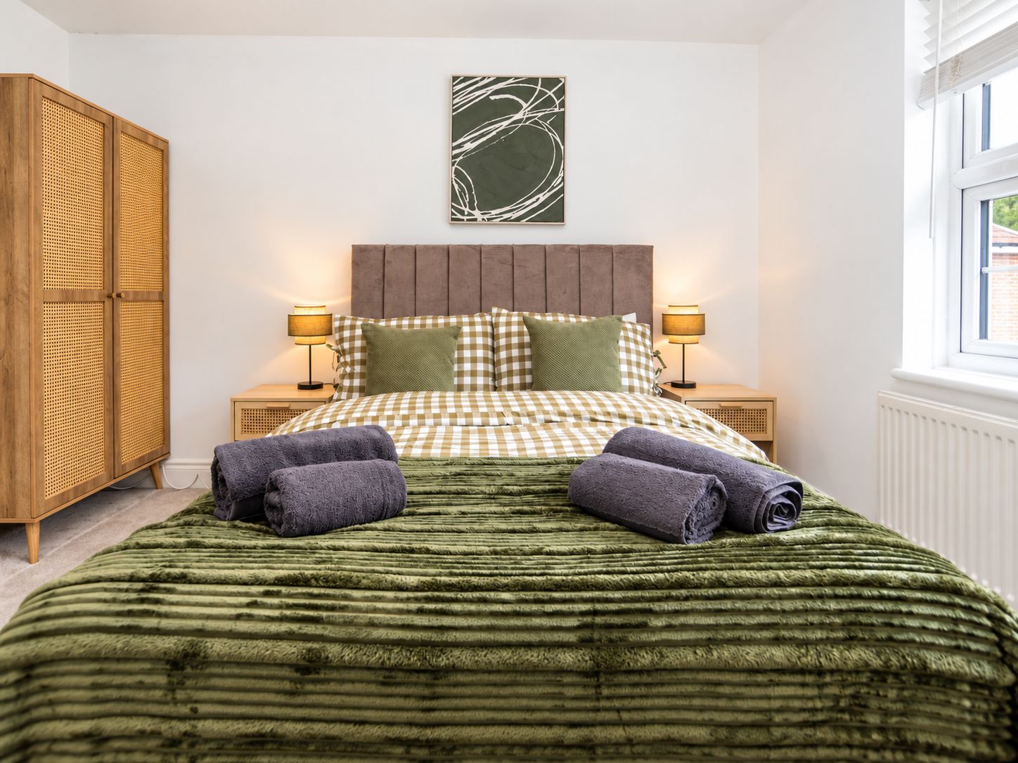 Ein Schlafzimmer mit kariertem Bettbezug, zwei Nachttischlampen, einem Holzschrank und einem Fenster mit Jalousien.
