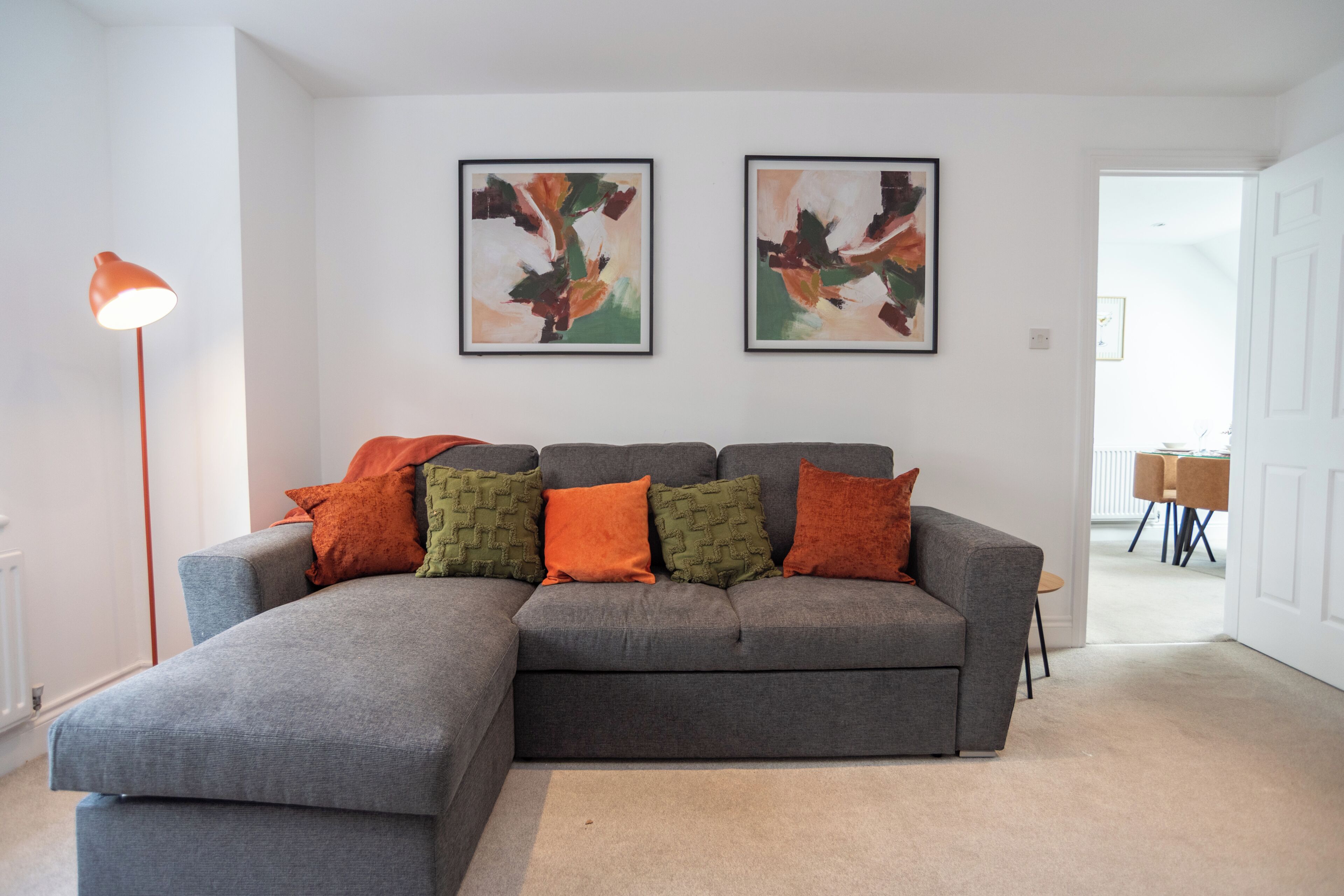Salón con un sofá seccional gris, varios cojines, dos cuadros abstractos, una lámpara de pie y vista a otra habitación.