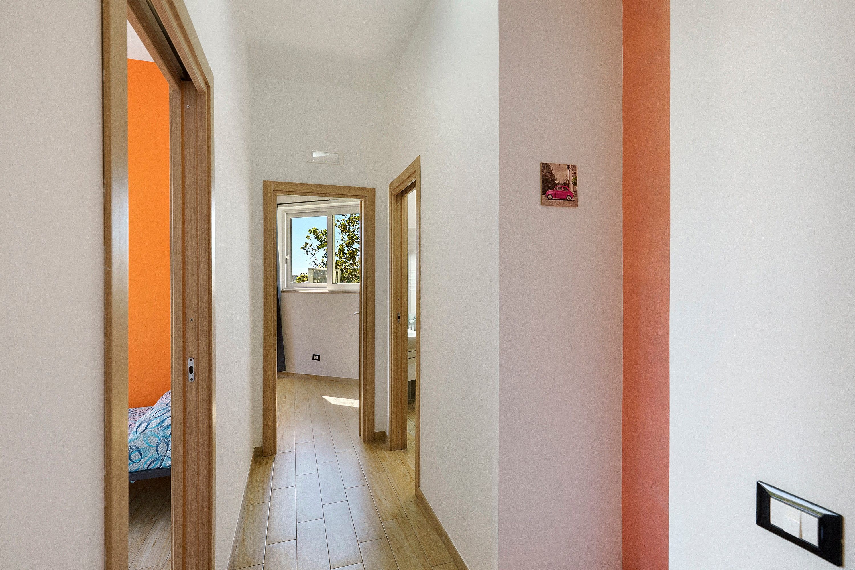 A narrow hallway with wooden flooring, white walls, and a door leading to a room with a patterned bedspread.