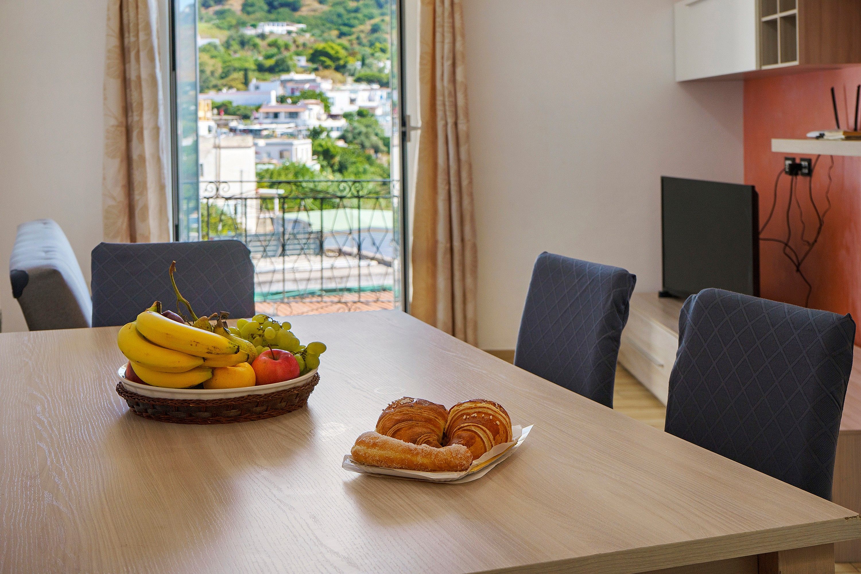 Un tavolo da pranzo con un cesto di frutta e pasticcini, vista su un quartiere residenziale attraverso una finestra e un televisore su un supporto.