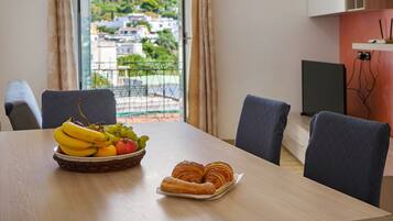 Un tavolo da pranzo con un cesto di frutta e pasticcini, vista su un quartiere residenziale attraverso una finestra e un televisore su un supporto.
