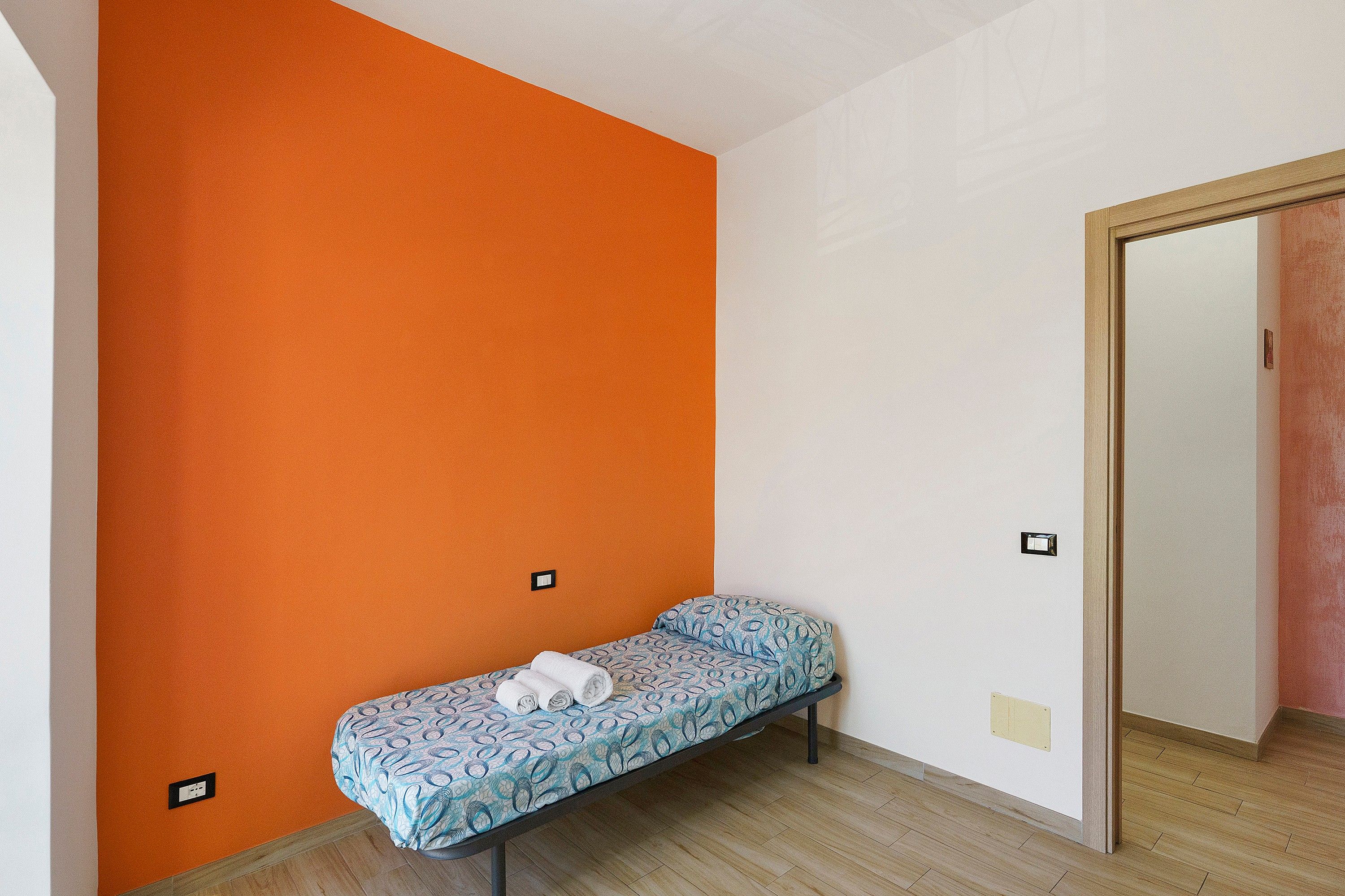 A small, single-bed room with an orange accent wall, a wooden floor, and a door leading to another room.