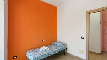 A small, single-bed room with an orange accent wall, a wooden floor, and a door leading to another room.