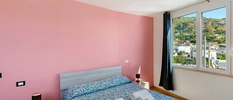 A bedroom with a bed, a nightstand, a lamp, and a view of a hillside.