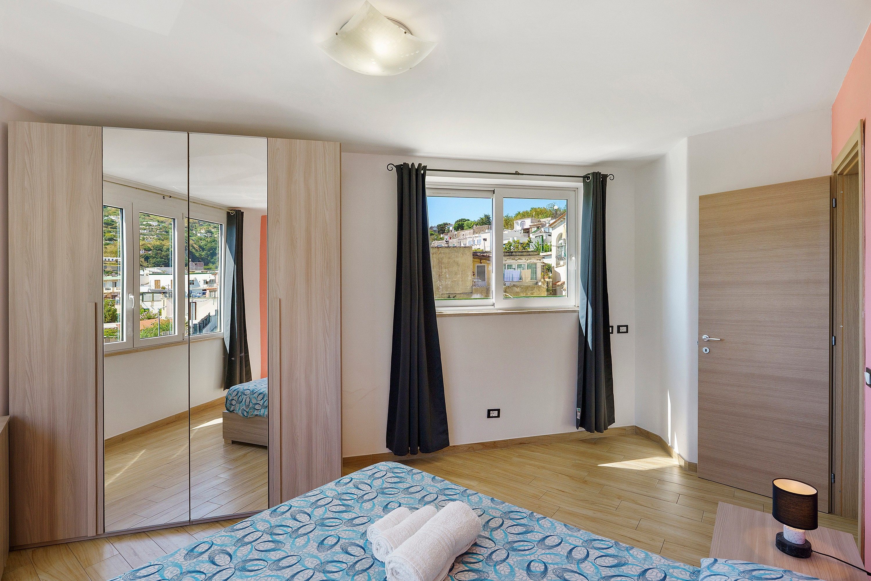 A bedroom with a bed, a wardrobe, a window with curtains, and a view of buildings outside.