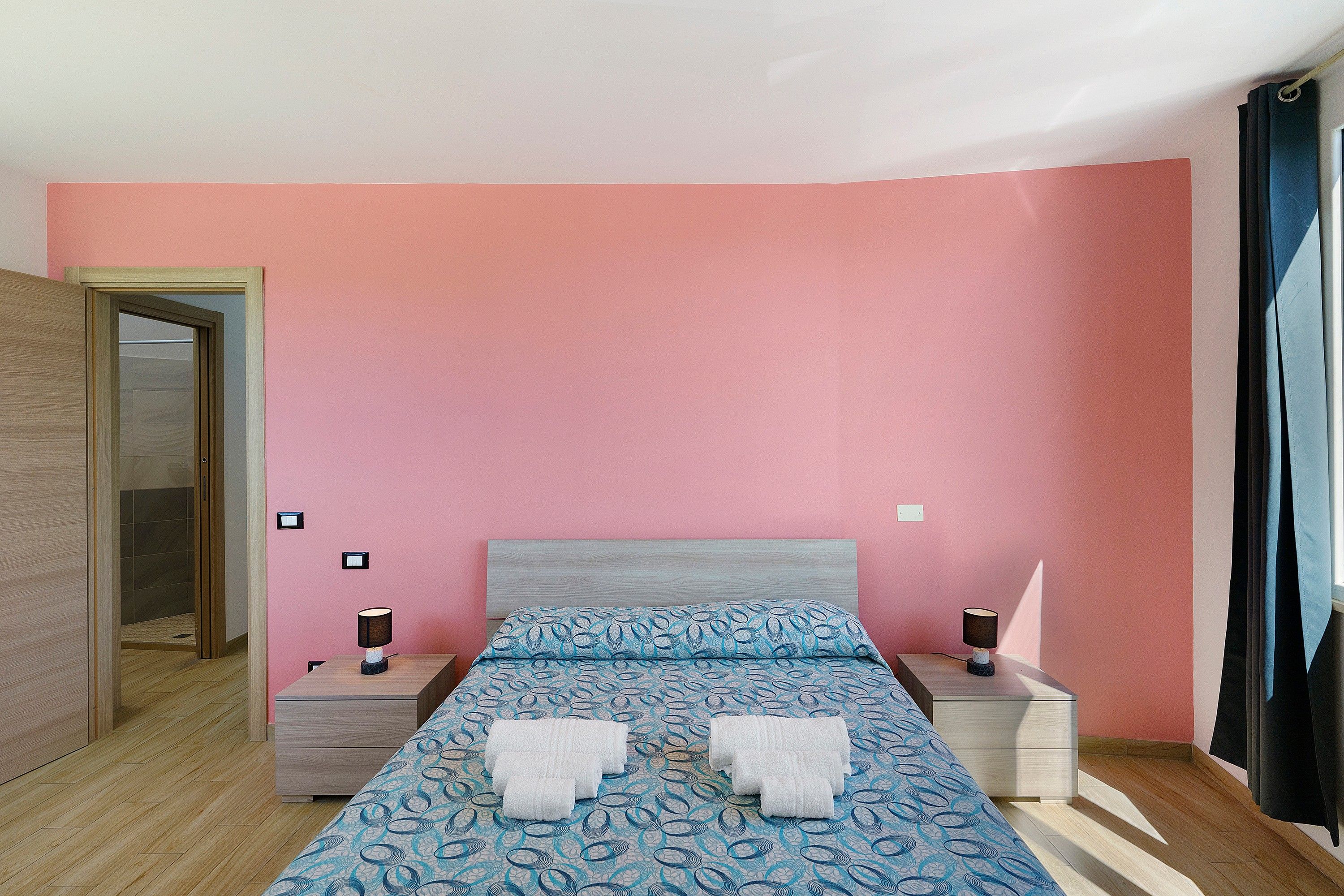 A bedroom with a pink wall, a bed with a patterned bedspread, two bedside tables with lamps, and a wooden wardrobe.