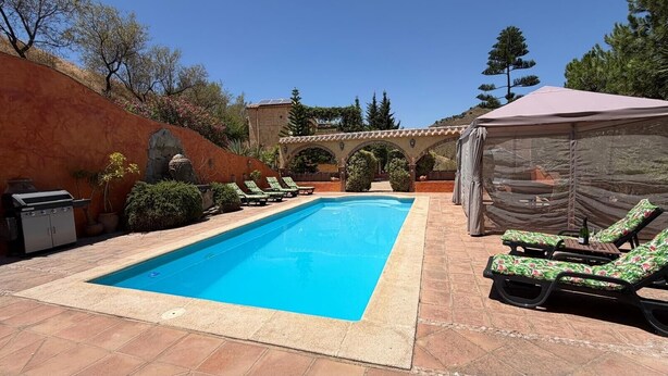 A rectangular swimming pool with a tiled deck, surrounded by lounge chairs and a covered seating area.