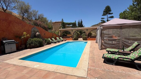 A rectangular swimming pool with a tiled deck, surrounded by lounge chairs and a covered seating area.