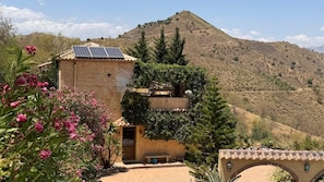 A building with a solar panel, surrounded by vegetation and a hilly landscape.