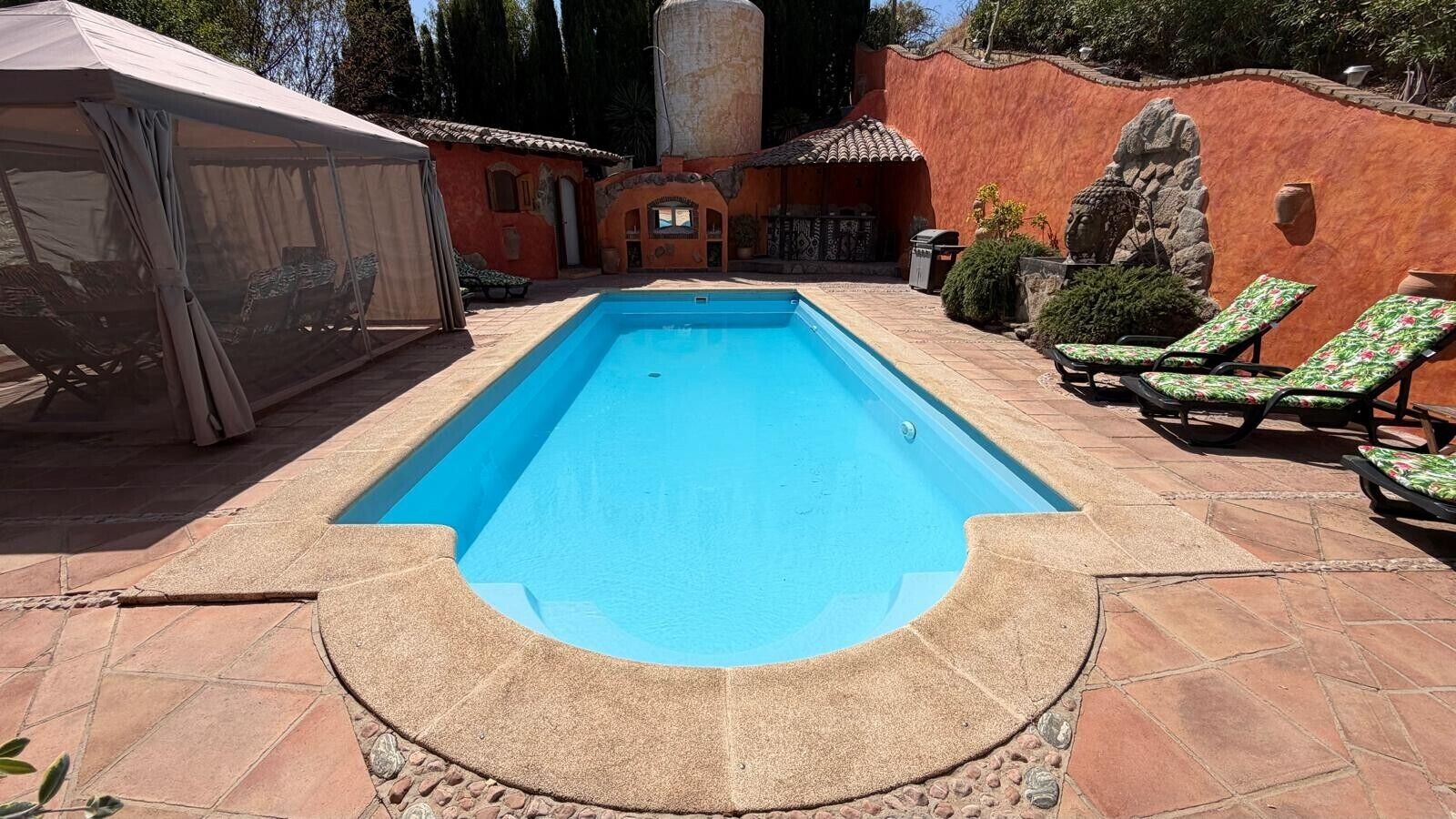 A rectangular swimming pool with a tiled deck, surrounded by lounge chairs and a covered seating area.