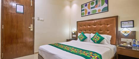 A hotel room with a large bed, a wooden door, a nightstand with a lamp, and a wall-mounted artwork.