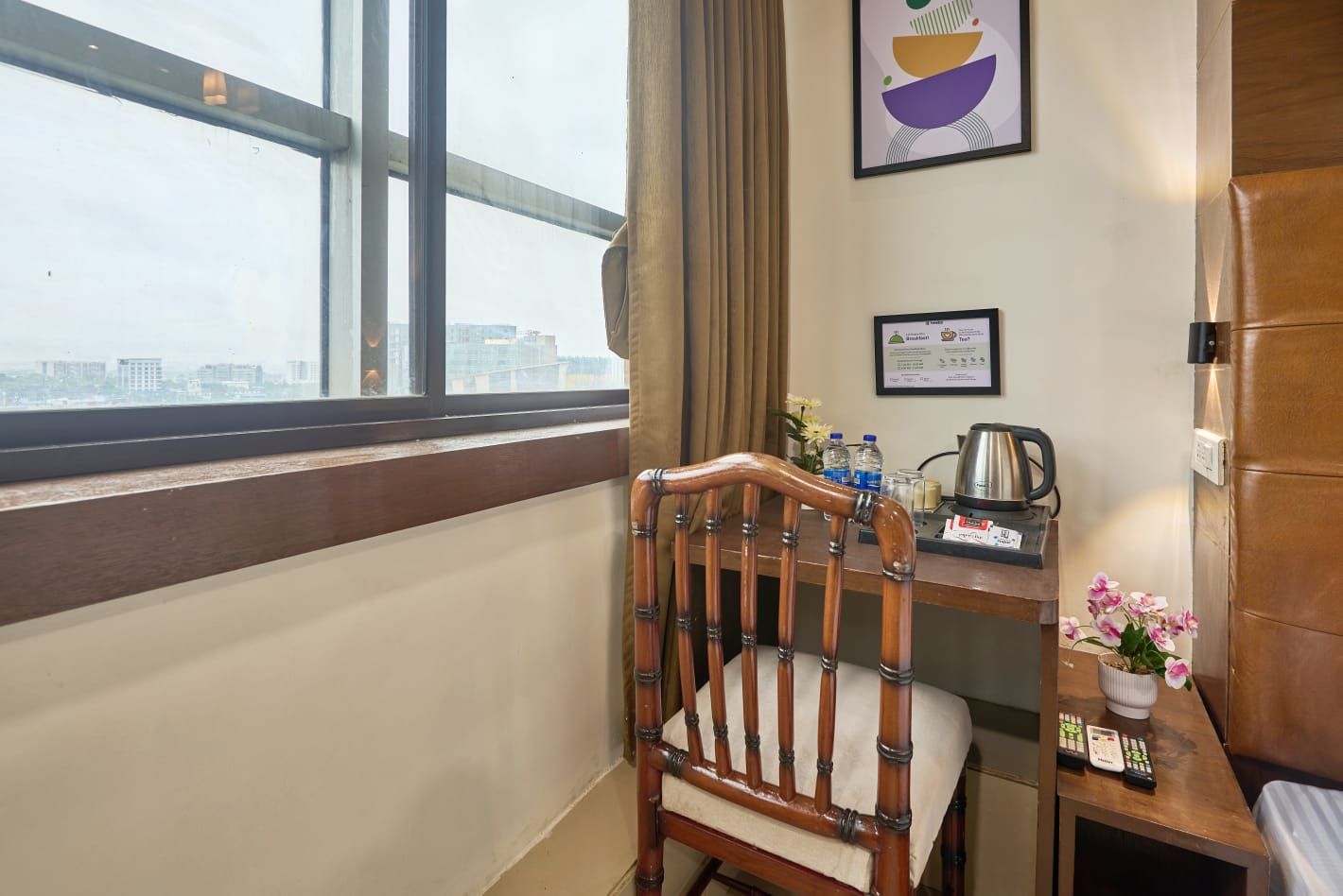 Habitación de hotel con una silla de madera, un escritorio con un hervidor, un certificado enmarcado y una ventana con vistas a la ciudad.