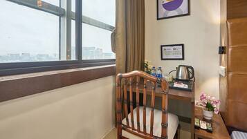 Habitación de hotel con una silla de madera, un escritorio con un hervidor, un certificado enmarcado y una ventana con vistas a la ciudad.