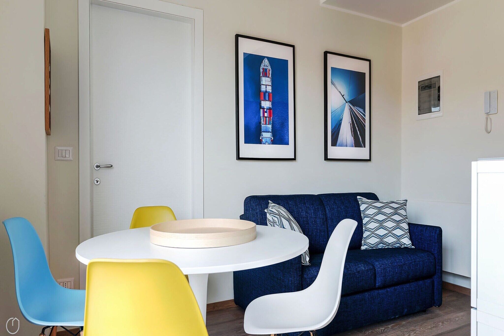 A room with a white door, a round table with a beige tray, a blue sofa with patterned pillows, and two chairs, one yellow and one white.