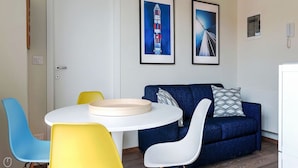 Quarto com porta branca, mesa redonda com bandeja bege, sofá azul com almofadas estampadas e duas cadeiras, uma amarela e outra branca.