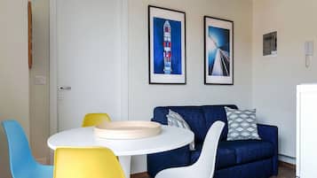 A room with a white door, a round table with a beige tray, a blue sofa with patterned pillows, and two chairs, one yellow and one white.