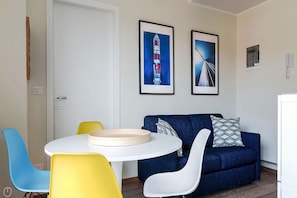 Une pièce avec une porte blanche, une table ronde sur laquelle repose un plateau beige, un canapé bleu agrémenté de coussins à motifs, et deux chaises, une jaune et une blanche.