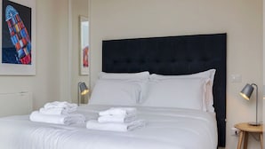 Cama bem arrumada com roupas de cama e toalhas brancas, uma cabeceira estofada preta e duas luminárias de mesa.
