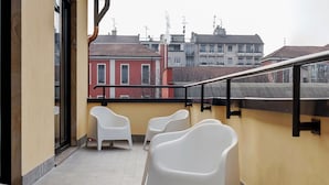 Uma varanda com cadeiras de plástico branco, piso de azulejos e vista para prédios ao fundo.