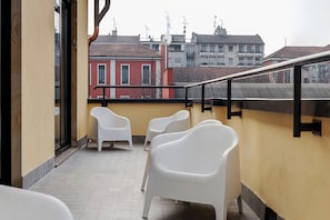 Un balcon avec des chaises en plastique blanches, un sol carrelé et une vue sur les bâtiments en arrière-plan.