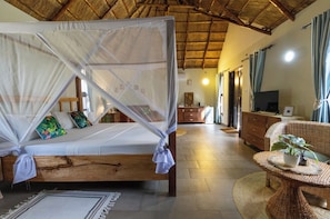 A room with a bed under a mosquito net, a wicker chair, a television, and a wooden cabinet. - Luxury Pool View Villa, Terrace & AC Zanzibar (Pwani Mchangani)