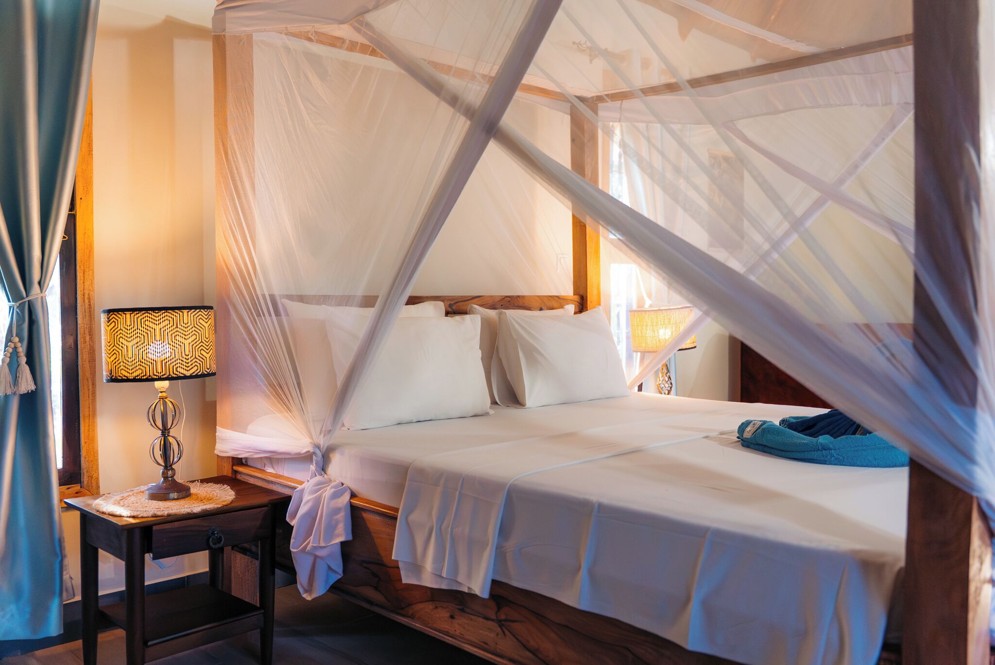 A room with a bed under a canopy, a bedside table with a lamp, and a window with curtains.