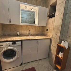 Une petite cuisine équipée d’un lave-linge, d’un évier et d’une étagère en bois sur laquelle est posé un rouleau de papier toilette.