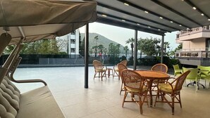 A covered outdoor seating area with wicker chairs and a cushioned bench, overlooking a pool and buildings.