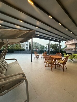 Un espace extérieur aménagé, abrité et doté de chaises en osier et d’un banc rembourré, offrant une vue sur une piscine et des bâtiments.