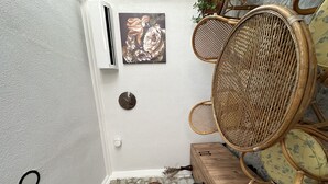 A corner of a room with a white air conditioning unit, a wall clock, a wicker chair, a small wooden table, and decorative woven items on the floor.