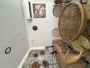 Un coin de pièce avec un climatiseur blanc, une horloge murale, une chaise en osier, une petite table en bois et des objets décoratifs en fibres tressées posés sur le sol.