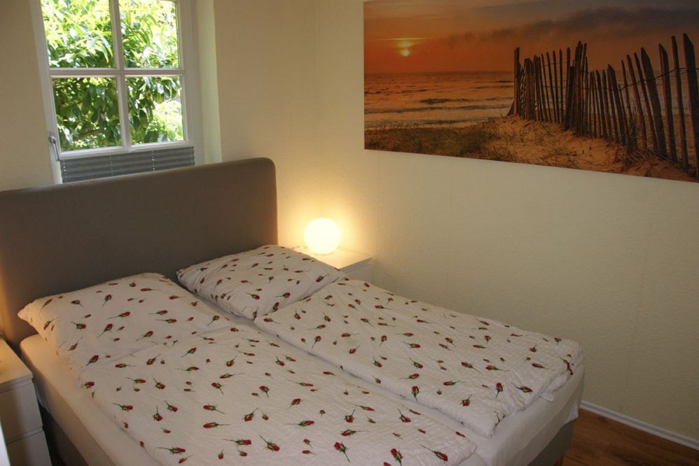 A bedroom with a bed, a window, and a beach sunset painting on the wall.