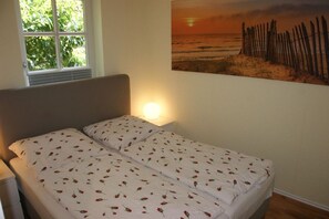 Une chambre avec un lit, une fenêtre et un tableau représentant un coucher de soleil sur la plage, accroché au mur.