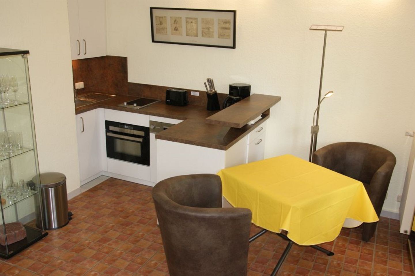 A compact kitchen with a dining area, including a table with a yellow tablecloth, two chairs, and a trash bin.