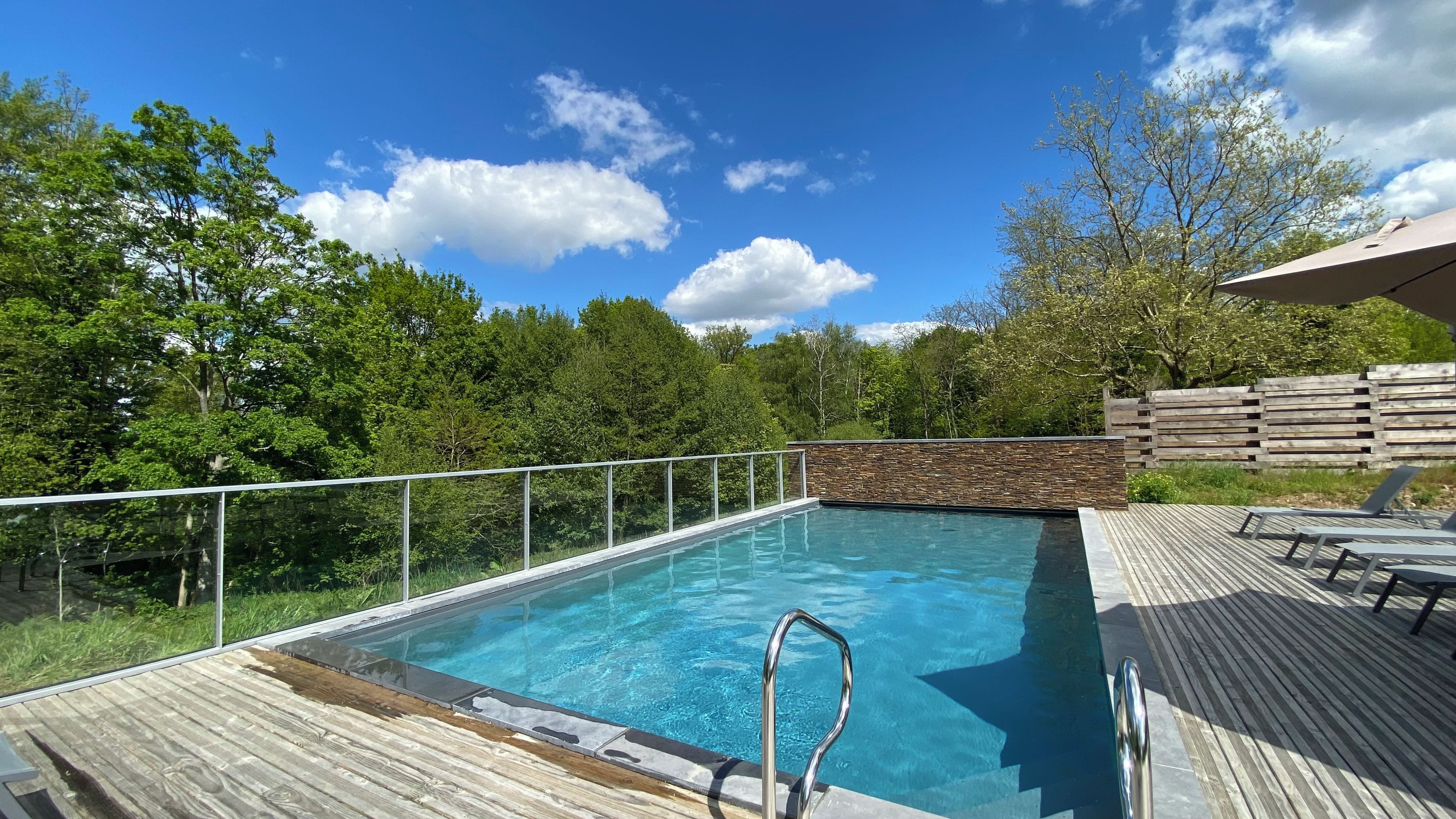 Una piscina rectangular con una escalera metálica, rodeada por una cubierta de madera y un muro de piedra.