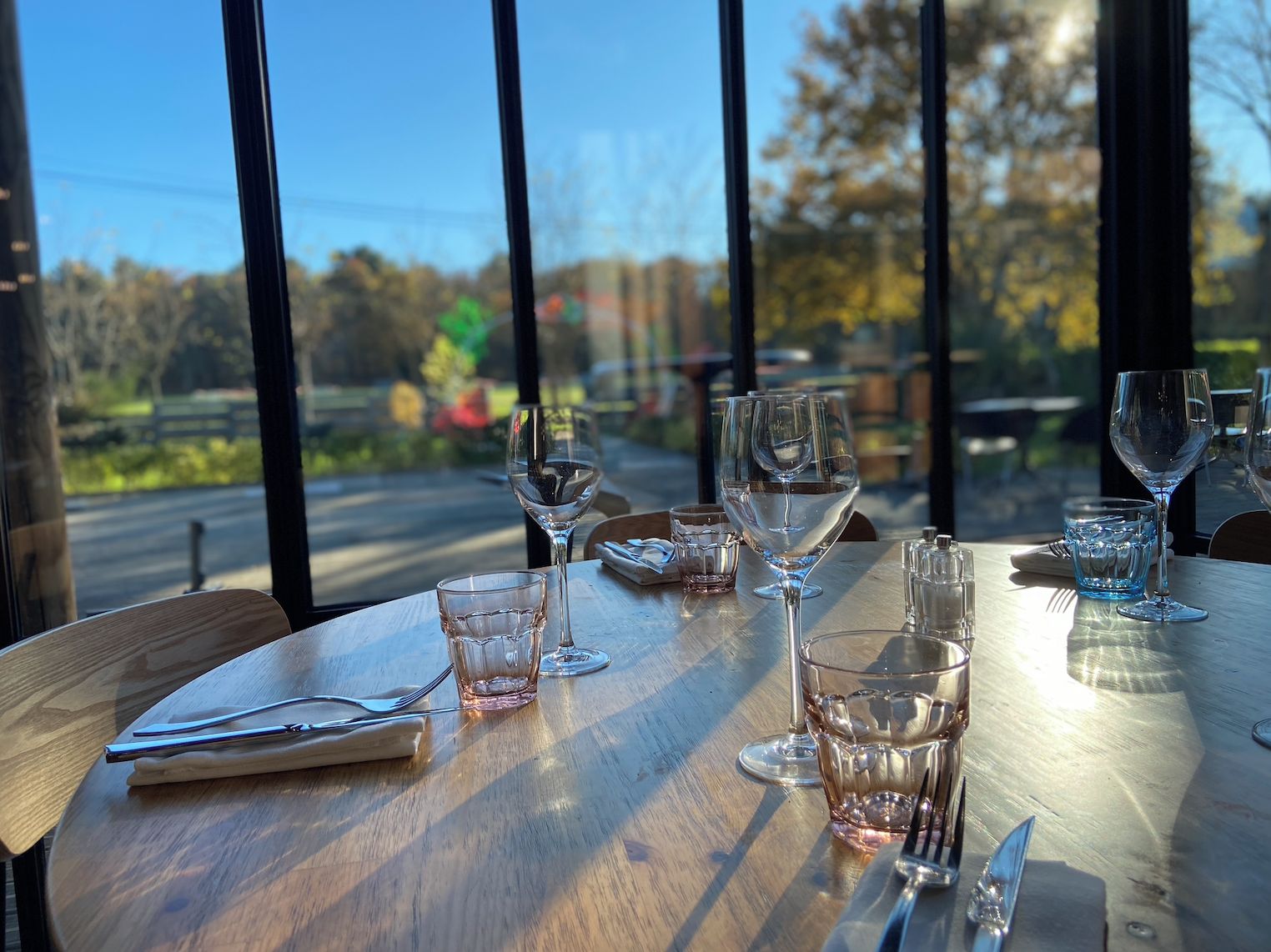 Una mesa de comedor con varias copas de vino y cubiertos, frente a una ventana con vistas a árboles y una carretera.