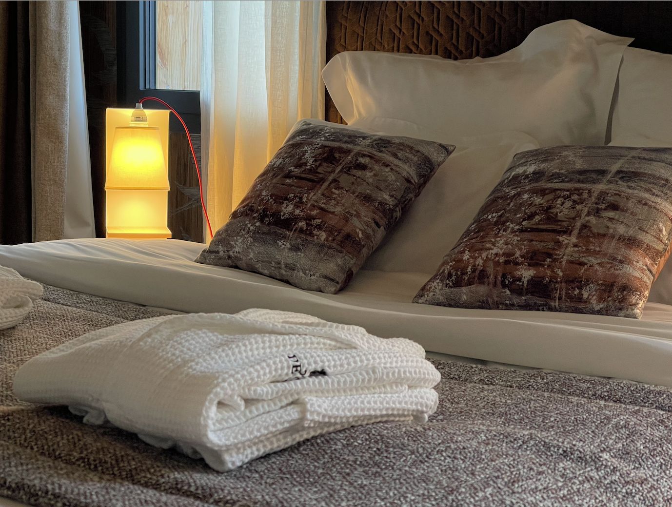 Una cama bien hecha con dos almohadas, una toalla blanca doblada y una lámpara de noche.