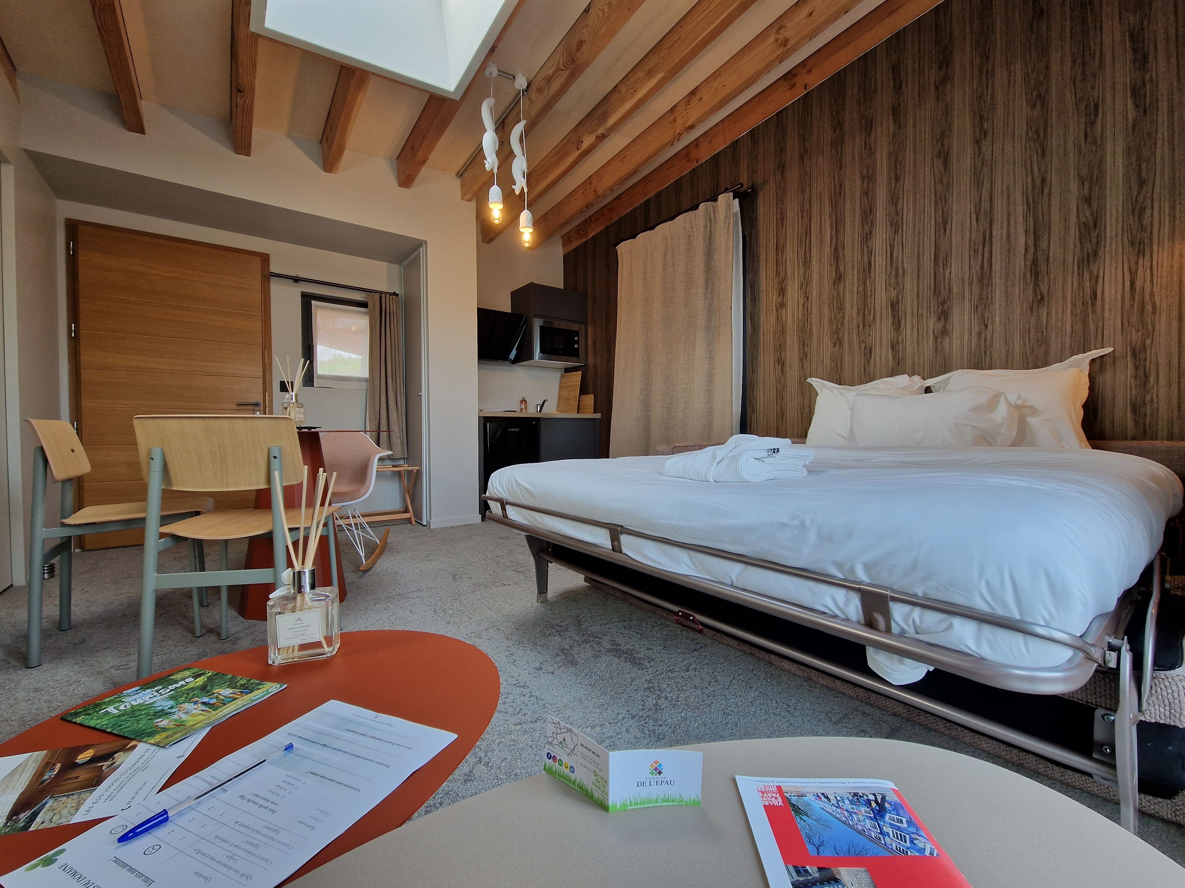 A hotel room with a bed, a desk with a chair, a wooden wardrobe, a microwave, and a ceiling with exposed beams.